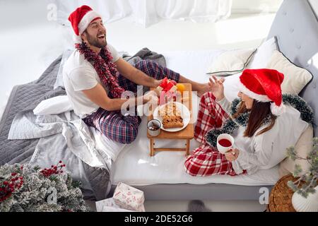 Junges Paar, das Spaß hat, lächelt, lacht, Frühstück im Bett isst, Geschenke gegen weihnachten austauscht, Weihnachtsmützen trägt Stockfoto