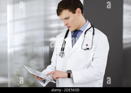 Ein junger, freundlicher Arzt überprüft einige Informationen auf seinem Computer-Tablet. Porträt eines Facharztes bei der Arbeit in einer Klinik. Medizinkonzept Stockfoto