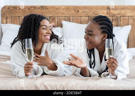 Glückliche Freundinnen in Bademänteln, die auf dem Bett liegen und Champagner trinken Stockfoto