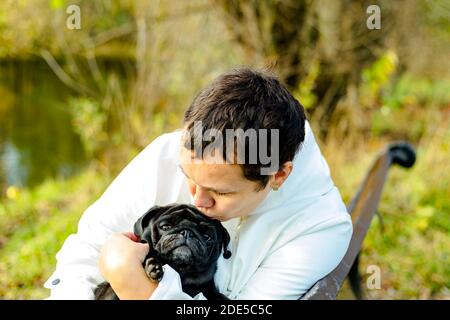 Frau mittleren Alters in weißer Jacke sitzt auf Parkbank und umarmt ihren schwarzen Pug Hund Stockfoto