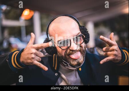 Portrait von glücklichen Hipster Mann trägt Brille, Gesichtsmaske, machen die Zunge und zeigt Teufel Dornen Geste Zeichen - Neue normale Lifestyle-Konzept mit mi Stockfoto