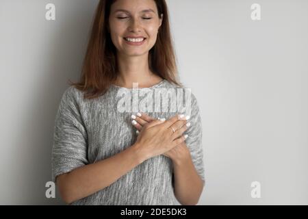 Lächelnde junge Frau hält gefaltete Hände auf der Brust und fühlt sich dankbar. Stockfoto