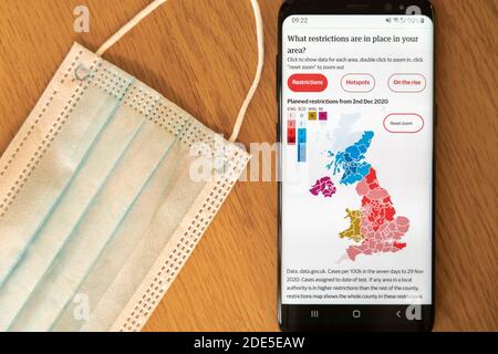 Geplante lokale Einschränkungen für Coronavirus Covid-19 in Großbritannien vom 2. Dezember 2020 auf einem Handy-Bildschirm mit Eine Gesichtsmaske im Hintergrund Stockfoto