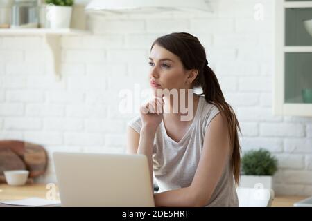 Verträumte nachdenkliche Frau, die am Tisch mit Laptop in der Küche sitzt Stockfoto