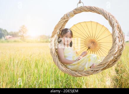 Glückliches Mädchen sitzt in Korbschaukel zwischen den goldenen Reisfeldern. Freizeit- und Erholungskonzept Stockfoto