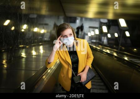 Junge Frau in einer gelben Jacke und einer Einwegmaske Fährt die Rolltreppe in der U-Bahn hoch Stockfoto