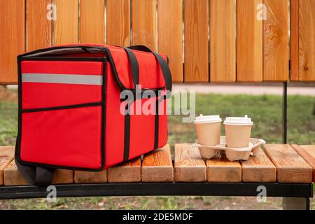 Große rote Tasche mit Aufträgen der Kunden des zeitgenössischen Cafés Oder Restaurant und zwei Pappgläser mit frischem heißen Kaffee Auf der hölzernen Bank im Park Stockfoto