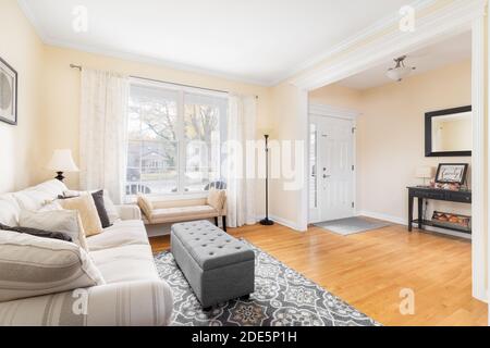 Ein helles, gemütliches Wohnzimmer mit Couch und Ottomane mit Blick auf ein großes Erkerfenster und Foyer. Stockfoto