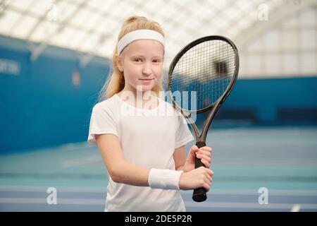 Nette blonde kleine Mädchen in weißen activewear hält Tennisschläger An der linken Schulter, während Sie vor der Kamera stehen stadionumgebung Stockfoto