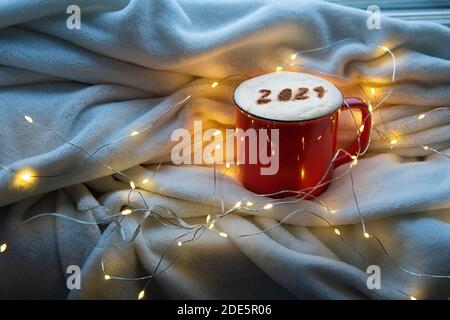 Rote Kaffeetasse mit der Nummer 2021 auf der schaumigen Cappuccino-Oberfläche auf dem kuscheligen karierten und weihnachtlichen Hintergrund. Blaues Nachtlicht. Frohes neues Jahr Stockfoto