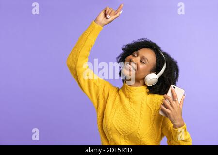 Energiegeladene Mixed Race Frau singt mit, lächelt breit, trägt kabellose Kopfhörer, hat gute Laune, isoliert auf violettem Hintergrund. Fröhliche dunkelhäutige Locke Stockfoto