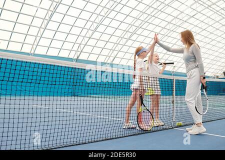 Glückliche Mädchen im Teenageralter in aktiven Jahren, die ihren hohe fünf gaben Junge Tennistrainer über Netz vor oder nach dem Training auf Modernes Stadion Stockfoto