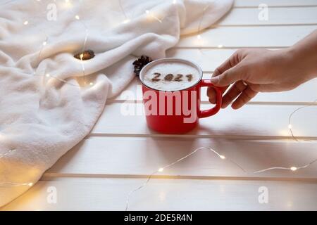 Rote Tasse Cappuccino mit der Nummer 2021 auf der schaumigen Oberfläche auf dem kuscheligen karierten und weihnachtlichen Lichtern Hintergrund. Die Hand nimmt eine Tasse Kaffee. Essenskunst Stockfoto