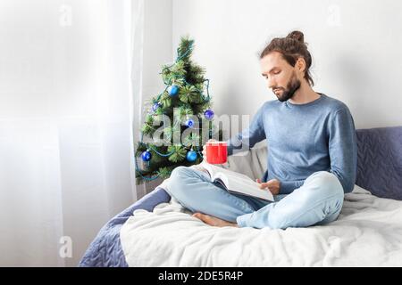 Junger bärtiger Mann sitzt auf dem Sofa auf der Decke und liest Buch und hält eine Tasse Kaffee in der Nähe von weihnachtsbaum zu Hause. Schön zu Hause im Winter und Stockfoto