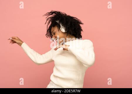 Energiegeladene Biracial Frau in weißem Pullover singen mit, trägt kabellose Kopfhörer, hat gute Laune, isoliert auf rosa Hintergrund. Glücklich dunkelhäutig gelockt Stockfoto