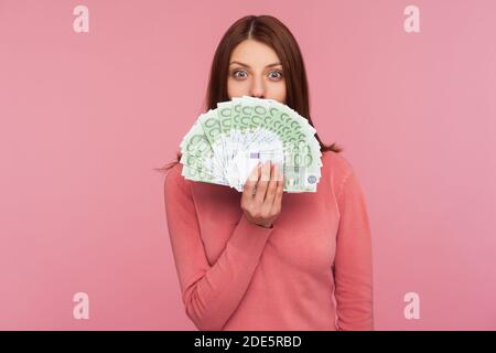 Verblüffte glückliche Frau mit braunen Haaren in rosa Pullover versteckt Gesicht hinter Fan von Euro-Banknoten, zinslose Barabhebung. Innenaufnahmen im Studio Isolate Stockfoto