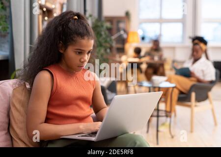 Serious African schulmädchen sitzt auf Couch im Wohnzimmer mit Laptop Auf den Knien und beim Blick auf das Display während des Online-Unterrichts Gegen ihre Familie Stockfoto