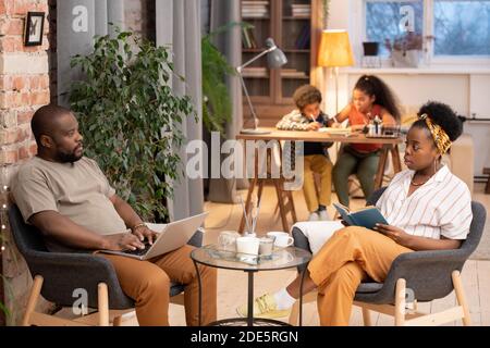 Junge afrikanische Frau mit offenem Buch und ihr Mann mit Laptop sitzt in grauen Sesseln am kleinen Tisch, während ihre Kinder machen Hausaufgaben Stockfoto