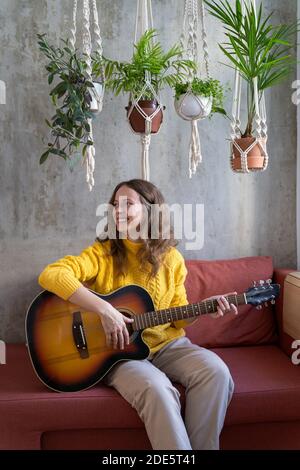 Lächelnde Frau Freiberuflerin in gelben Pullover, ruhend, sitzend auf Couch unter Baumwolle Macrame Pflanze Kleiderbügel mit Zimmerpflanzen, spielt die Gitarre. Hobby, han Stockfoto