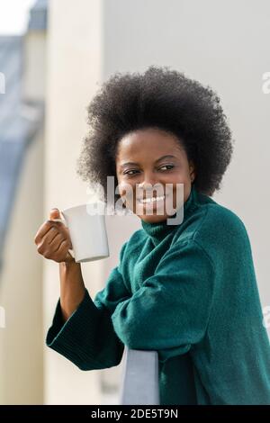 Lächelnde junge afroamerikanische Biracial Frau genießt eine Tasse Tee oder Kaffee, tragen übergroße grüne Strickpullover, wegschauen, auf dem Balkon stehen Stockfoto