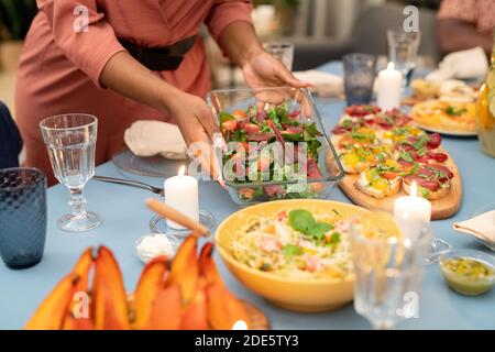 Hände der jungen afrikanischen Frau setzen Glas Schüssel mit frisch Gemüsesalat auf servierten festlichen Tisch mit hausgemachten Sandwiches und Pasta Stockfoto