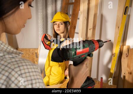 Zwei Mädchen in einem Werkzeug- und Holzlager präsentieren sie eine Handsäge für Holz und einen elektrischen Schraubendreher. Stockfoto