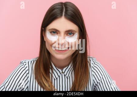 Positive schöne Frau mit braunen Haaren in gestreiftem Hemd mit Augenflecken Blick auf die Kamera mit toothy Lächeln, Schönheitsverfahren. Innenaufnahme im Studio Stockfoto