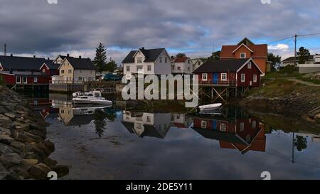 Henningsvær, Austvågøya, Lofoten, Norwegen - 08-29-2020: Traditionelle Holzhäuser und rote Rorbu (Fischerhaus auf Stelzen) mit Anlegeboot. Stockfoto