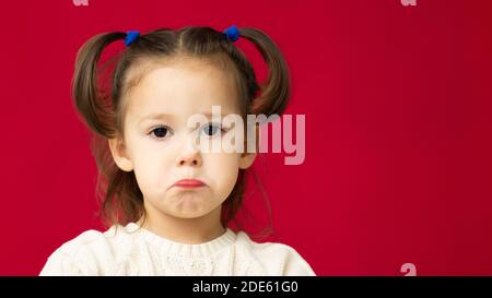 Das Gesicht eines traurigen kleinen niedlichen Mädchens 2-4 in einem weißen Strickkleid auf rotem Hintergrund. Stockfoto