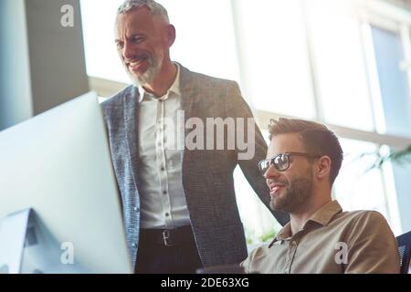 Fröhlicher reifer Geschäftsmann, männlicher Chef erklärt etwas zu jungen Kollegen, Blick auf PC-Bildschirm und lächelt während der Zusammenarbeit in Coworking Stockfoto