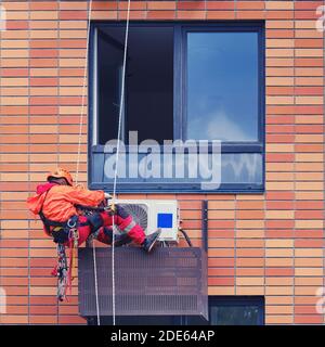 Ein männlicher Arbeiter in Uniform mit einem Sicherheitsseil Eine Klimaanlage in einer Nische unter dem Fenster Stockfoto
