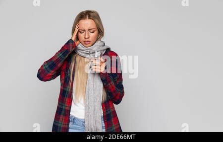 Frau im karierten Hemd, gewickelter Schal, der Kopfschmerzen hat und Glas mit Wasser hält, ihren Tempel berührt und die geschlossenen Augen isoliert. Grippesaison Stockfoto