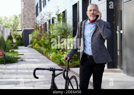Stilvoller, reifer Geschäftsmann, der beim Telefonieren mit dem Fahrrad tagsüber im Freien steht, wegschaut Stockfoto