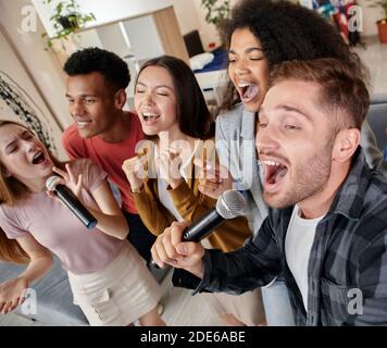 Stellen Sie sich dem Lied. Enthusiastische multikulturelle Freunde, die mit Mikrofon singen, während sie zu Hause Karaoke spielen und in der modernen Wohnung stehen Stockfoto