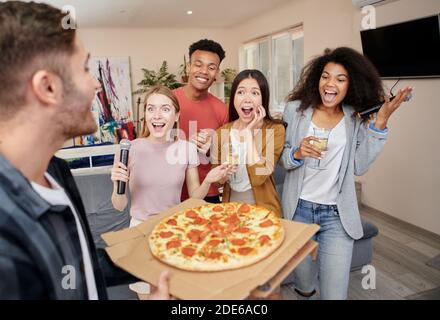 Pizza-Zeit. Aufgeregt multikulturelle Freunde mit Pizza und Karaoke-Party, singen mit Mikrofon, während zusammen in der modernen Wohnung stehen Stockfoto