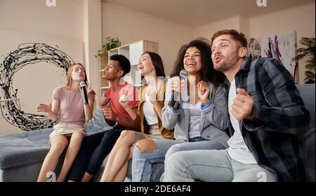 Singen Sie einfach. Fröhliche multikulturelle Freunde, die enthusiastisch aussehen, während sie zu Hause Karaoke spielen, mit Mikrofon singen und drinnen auf der Couch sitzen Stockfoto