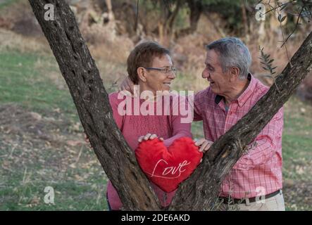 Glücklicher älterer Mann und Frau mit rotem Herzen und Wort Liebt einander, während ihr in der Nähe von Bäumen im grünen Garten steht Stockfoto