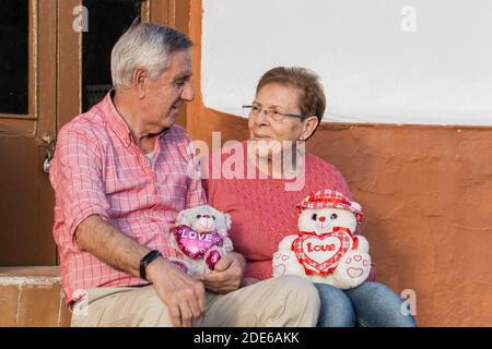 Positive ältere Mann und Frau in Casual tragen niedlich Spielzeug mit Herzen und einander mit Liebe betrachten Während der Feier des Valentinstag Stockfoto