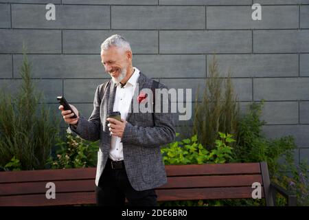 Soziale Medien werden überprüft. Portrait von fröhlichen, selbstbewussten Geschäftsmann im mittleren Alter in stilvollen Anzug lächelnd, während mit seinem Smartphone, im Freien stehend Stockfoto