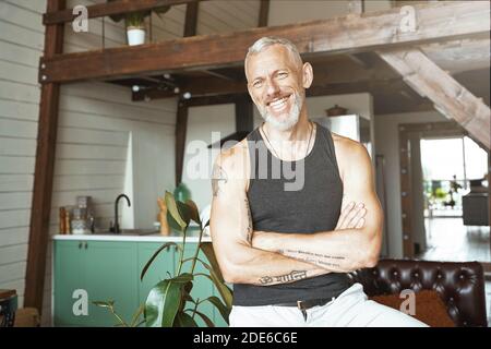 Porträt eines gutaussehenden fröhlichen kaukasischen reifen Mannes mit gekreuzten Armen, lächelnd in der Kamera, während er in seiner modernen Wohnung steht Stockfoto