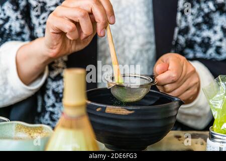 Teemeisterin Motoko Dobashi siebt Matcha-Pulver durch ein feines Sieb mit einem Bambislöffel (Chashaku) in eine dunkle Teeschale während einer japanischen Teezeremonie im Teeladen ANMO in Düsseldorf, Deutschland. Ein Bambus-Teewhipper (Chasen) und eine Dose mit zusätzlichem Matcha-Pulver sind bereitgelegt. Das sind vermutlich die Hände von . Stockfoto