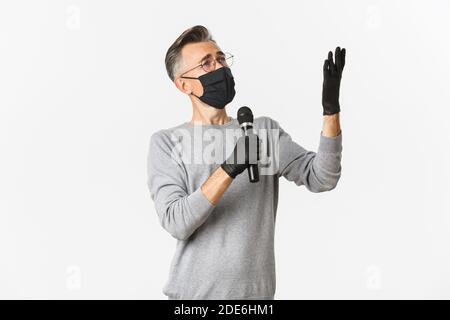 Konzept von Coronavirus, Lebensstil und Quarantäne. Porträt eines charismatischen Mannes mittleren Alters in medizinischer Maske und Handschuhen, singende Serenade im Mikrofon Stockfoto