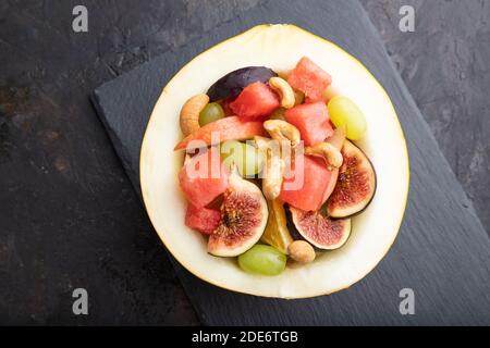 Vegetarischer Obstsalat aus Wassermelone, Trauben, Feigen, Birne, Orange, Cashew auf Schiefertafel auf schwarzem Betongrund. Draufsicht, flach liegend, Nahaufnahme. Stockfoto