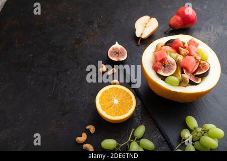 Vegetarischer Obstsalat aus Wassermelone, Trauben, Feigen, Birne, Orange, Cashew auf Schiefertafel auf schwarzem Betongrund. Seitenansicht, Kopierbereich. Stockfoto