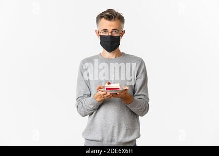 Konzept von Coronavirus, Quarantäne und Urlaub. Bild von traurigen Mann mittleren Alters in schwarzer medizinischer Maske und Brille, hält Geburtstagskuchen mit brennenden Kerze Stockfoto