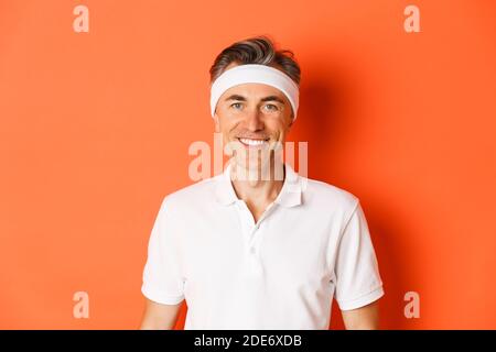 Nahaufnahme eines gutaussehenden Sportlers mittleren Alters, mit Stirnband und weißem T-Shirt, lächelnd glücklich, stehend auf orangefarbenem Hintergrund Stockfoto