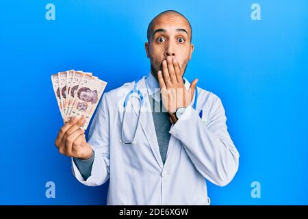 Hispanische Erwachsene Arzt Mann trägt medizinische Uniform hält 500 mexikanische Pesos über den Mund mit der Hand, schockiert und Angst vor Fehler. Überrascht Expres Stockfoto