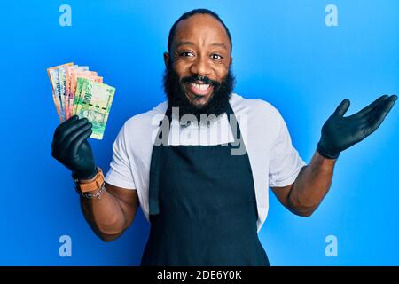 Junger afroamerikanischer Mann mit professioneller Schürze, der Südafrika hält rand Banknoten feiern Leistung mit glücklichen Lächeln und Gewinner expressi Stockfoto