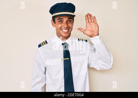 Junger hispanischer Mann trägt Flugzeug Pilot Uniform waiving sagen Hallo glücklich und lächelnd, freundliche Willkommensgeste Stockfoto