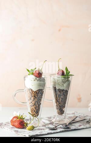 Zwei Gläser gesundes Frühstück mit Müsli und griechischem Joghurt, dekoriert mit Minze und roher Erdbeere, serviert auf weißem Holzhintergrund. Stockfoto